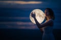 Tender image of a girl. Female magic. Beautiful attractive girl on a night beach with sand hugs the moon, art photo. On a dark