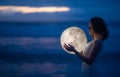 Tender image of a girl. Female magic. Beautiful attractive girl on a night beach with sand hugs the moon, art photo. On a dark