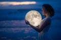 Tender image of a girl. Female magic. Beautiful attractive girl on a night beach with sand hugs the moon, art photo. On a dark