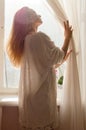 Tender pretty young blond woman standing near window at home or hotel room and luxuriating in the sun light flares Royalty Free Stock Photo