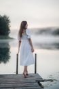 Tender romanitc sentimental female lady in the morning on a wooden pier near the misty river in a white dress Royalty Free Stock Photo