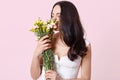 Tender sweet brunette young lady standing straight, holding beautiful flowers in both hands, feeling their spring smell, looks Royalty Free Stock Photo