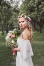 tender young bride in floral wreath and wedding dress holding bouquet of flowers and looking at camera outdoors Royalty Free Stock Photo