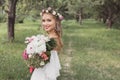 tender young bride in floral wreath and wedding dress holding bouquet of flowers and smiling at camera outdoors Royalty Free Stock Photo