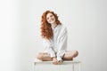 Tender young redhead girl in shirt smiling looking at camera sitting on table posing over white background. Royalty Free Stock Photo