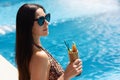 Tenned beautiful woman in leopard print swimming suit and black sunlasses sitting in swimming poolside edge with cocktail in hand