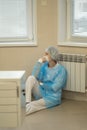 A tense female doctor sits tiredly on the hospital floor wearing a face mask and blue uniform.