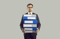 Tense male office worker with stressed expression holding stack of heavy folders on gray background.