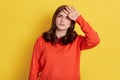 Tensed woman touching her head and looking directly at camera with tired painful look dresses orange jumper isolated over yellow