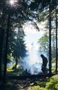 Tent among fir trees under the rays of sun breaking through clouds on cold day. Guy making fire and smoke flows up Royalty Free Stock Photo