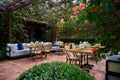 Terrace with chairs and tables setting with candles and vases with flowers, green bush on the foreground