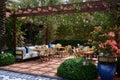 Terrace with blue couches, chairs and tables setting with candles and vases with flowers, green bushes on the foreground