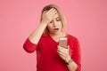Terrfied blonde young woman stares at camera of mobile phone, keeps hand on forehead, reads bad news on internet website, isolated