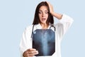 Terrific female doctor in white robe examines X ray picture of spine, holds hand on head, realizes that patient has scoliosis, iso Royalty Free Stock Photo