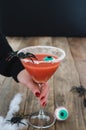 Terrifying red cocktail with spider on black background and wooden Halloween table with decoration, woman hand holding cup.