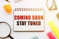 The text coming soon stay tuned in a notebook on a white table with office tools Royalty Free Stock Photo