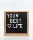 Text in english spelling `Your Best Life` with a heart on black felt board in a wooden frame. Letter Board on white background.