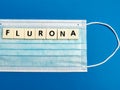 Text flurona made of square letter tiles with medical mask against blue background.