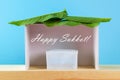 Text of Happy Sukkot. A hut made of paper covered with leaves on a blue background. Postcard, congratulations.