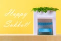 Text of Happy Sukkot. A hut made of paper covered with leaves on a yellow background. Postcard, congratulations.