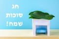 The text in Hebrew is Happy Sukkot. A hut made of paper covered with leaves on a blue background.