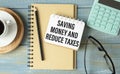 Text SAVING MONEY AND REDUCE TAXES on card on office desk with laptop, calculator, chart, clipboard, glasses, pen.