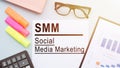 The text SMM - SOCIAL MEDIA MARKETING, on office desk with calculator, markers, glasses and financial charts