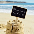 Text summer is here in a signboard topping a sandcastle Royalty Free Stock Photo