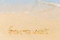 Text on a sunny beach. The word Forever written by hand in the sand, washed away by the sea wave. The concept of irony