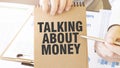 Text TALKING ABOUT MONEY on brown paper notepad in businessman hands on the table with diagram. Business concept Royalty Free Stock Photo