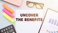 The text - UNCOVER THE BENEFITS on office desk with calculator, markers, glasses and financial charts