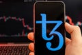 Tezos XTZ cryptocurrency logo on the screen of smartphone in mans hand with downtrend on the chart on a red light