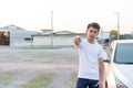 A Thai man in white shirt is pointing to camera in front of his silver car in the evening light