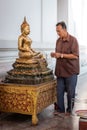 Thailand; Jan 2020: Buddhist man sticking gold leaves on golden Buddha statue, man praying in thankful pose. Sitting Buddha in