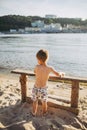 The theme is a child and summer beach vacation. A small Caucasian boy climbs up on a wooden bench on the river bank in a sunny sum