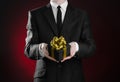Theme holidays and gifts: a man in a black suit holds exclusive gift wrapped in a black box with gold ribbon and bow on a dark red Royalty Free Stock Photo