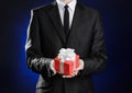 Theme holidays and gifts: a man in a black suit holds exclusive gift wrapped in red box with white ribbon and bow on a dark blue Royalty Free Stock Photo