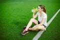 Theme sport and health. Beautiful young girl sitting resting on green grass, artificial turf stadium resting thirsty drink bottle