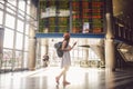 Theme travel and tranosport. Beautiful young caucasian woman in dress and backpack standing inside train station or terminal looki