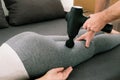 A therapist massages a woman's legs with a massage percussion device in her home. Percussion therapy of athlete's