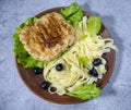 There is a clay plate with food on the table. On a plate, schnitzel with noodles. The food is decorated with salad and olives.