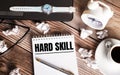 There is a cup of coffee on a wooden table, a clock, glasses and a notebook with the words HARD SKILL. Business concept