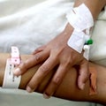 There is a middle-aged woman sore hand with a close-up infusion set and a wristband on a white hospital bed sheet.
