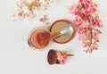 There White and Pink Branches of Chestnut Tree,Bronze Powder with Mirrow and Two Make Up Brushes are on White Table,Top View.Tone