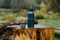 Thermos and aluminum hot drink mug with rising steam outdoors. Camping vacuum flask and iron cup standing on tree stump