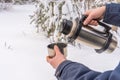 Thermos with hot tea or coffee in human hands against winter snow covered forest. Man pours drink into mug in cloudy winter day. Royalty Free Stock Photo