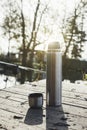 Thermos with hot tea, coffee stands on a wooden walkway in the sun on a pond or a small lake. Travel alone, outdoor recreation,