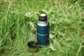 Thermos and a mug with a hot drink standing on the grass outdoors, top view. Steam rising from a steel cup