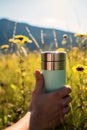 A thermos mug with hot tea in hand on a blurred background of a yellow-green mountain field with flowers in summer. Cup of coffee