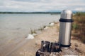 Thermos vacuum flask with hot tea and metal cups by summer lake. Camping, traveling and tourism concept